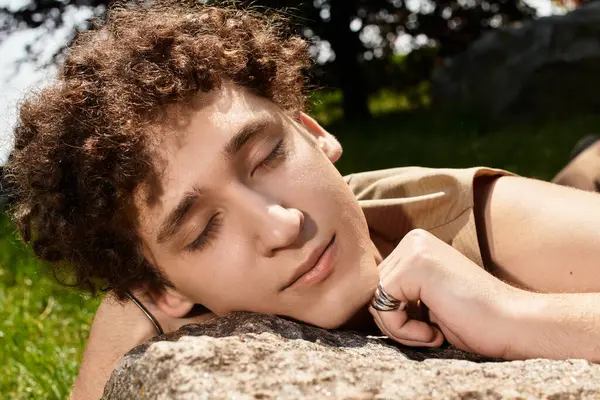A young man with curly hair relaxes on a rock in a sunny park, embracing summer warmth and style. — Stock Photo