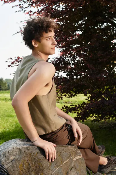 Curly-haired young man enjoys the outdoors while sitting on a rock surrounded by nature. — Stock Photo