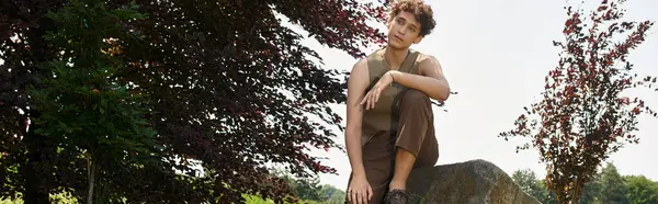 A young man with curly hair enjoys a sunny day in a lush green park, showcasing his stylish outfit, banner — Stock Photo