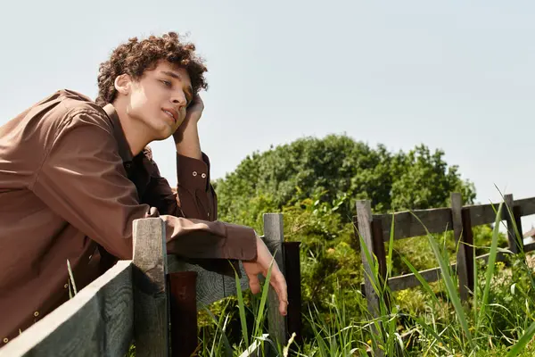 Curly-haired young man relaxes by a wooden fence in a lush park on a sunny day. — Stock Photo