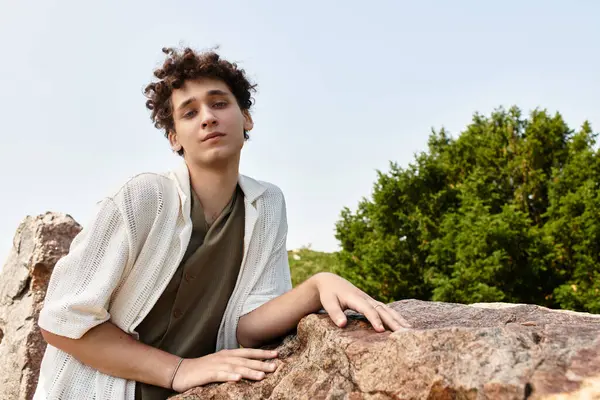 Young man with curly hair relaxes outdoors in a fashionable summer outfit amidst lush greenery. — Stock Photo