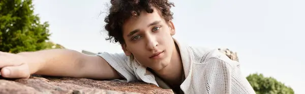 Curly-haired young man relaxes on a rock, soaking up the sunshine in a beautiful park, banner — Stock Photo