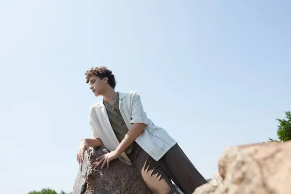 Curly-haired young man relaxes against a rock, soaking in the summer vibes in a lush park. — Stock Photo