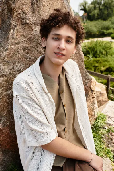 Curly-haired young man relaxes on a rock surrounded by lush greenery in the park. — Stock Photo