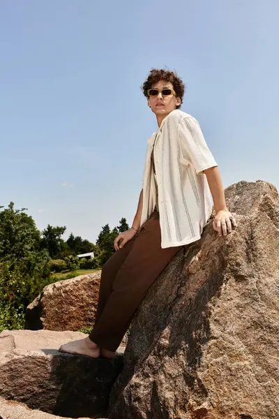 A young man with curly hair relaxes on a rock in a lush park, showcasing his trendy summer outfit. — Stock Photo