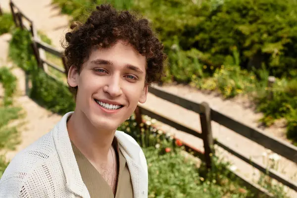 A young man with curly hair relaxes in a park, wearing a trendy summer outfit and smiling. — Stock Photo