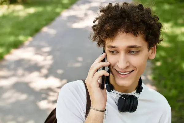 A young man with curly hair smiles while chatting on his phone in a lush green park. — Stock Photo