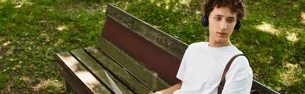 Curly-haired young man relaxes on a park bench, listening to music and soaking up the sun, banner — Stock Photo