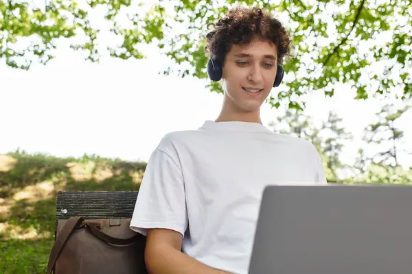 Curly-haired youth relaxes outdoors, focused on his laptop in a sunny green park. — Stock Photo