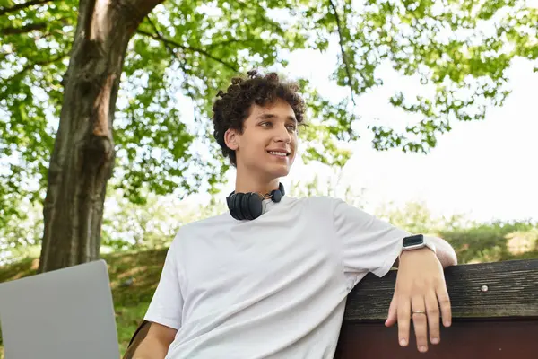 A young man with curly hair enjoys the warm summer air in a lush green park, feeling relaxed. — Stock Photo