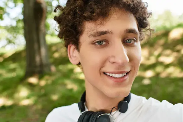 A young man with curly hair smiles while spending time outdoors in a lush green park. — Stock Photo