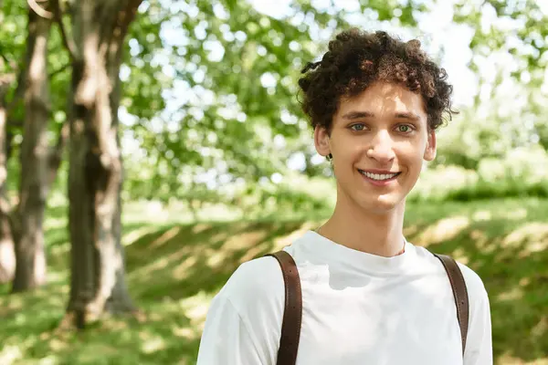 A young man with curly hair smiles warmly while relaxing in a sunny green park filled with trees. — Stock Photo
