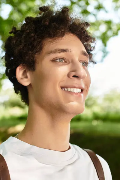 A young man with curly hair smiles brightly while soaking in the sun at a lush green park. — Stock Photo
