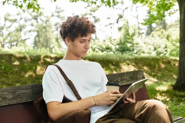 A young man with curly hair relaxes in a green park while working on his tablet and sipping coffee. — Stock Photo