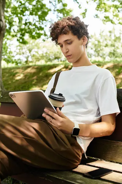 A young man with curly hair sits on a bench in a green park, focused on his tablet and coffee. — Stock Photo