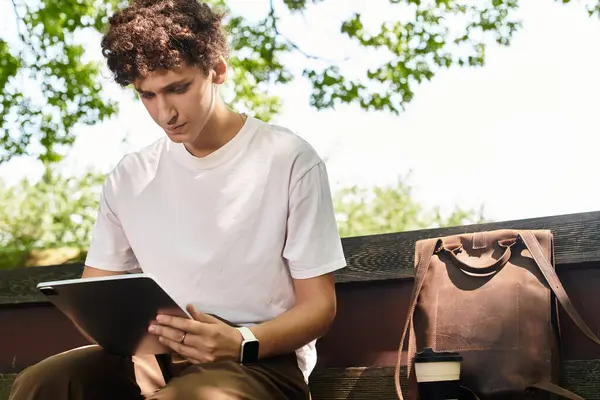 A young man with curly hair sits on a bench in a green park, focused on his tablet, relaxed. — Stock Photo