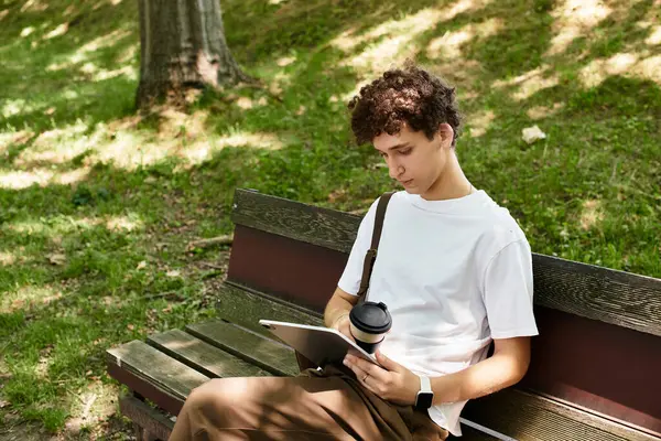 A young man with curly hair sits on a bench in a sunny park, savoring his coffee and relaxing. — Stock Photo