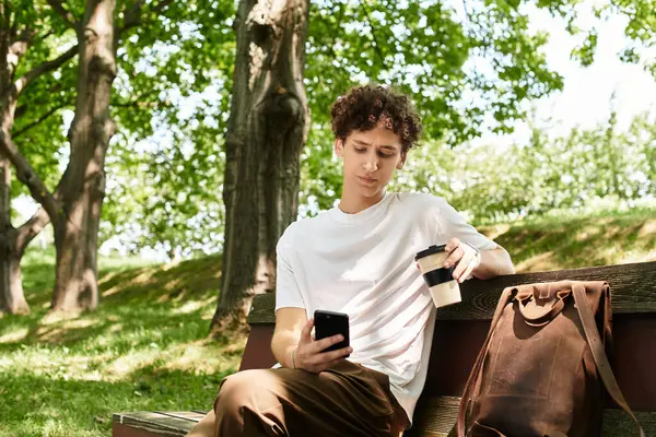 A young man with curly hair relaxes in a green park, sipping coffee and using his phone. — Stock Photo