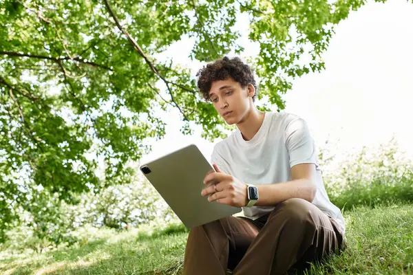 Curly-haired young man relaxes on the grass, engaged with his tablet in a vibrant park. — Stock Photo
