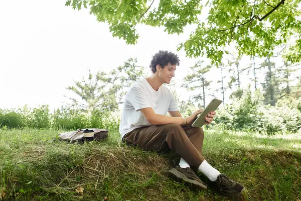 Curly-haired youth in a white t-shirt smiles while using a tablet in a sunny green park — Stock Photo