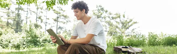 Relaxing under the sun, a young man with curly hair reads intently while surrounded by greenery, banner — Stock Photo