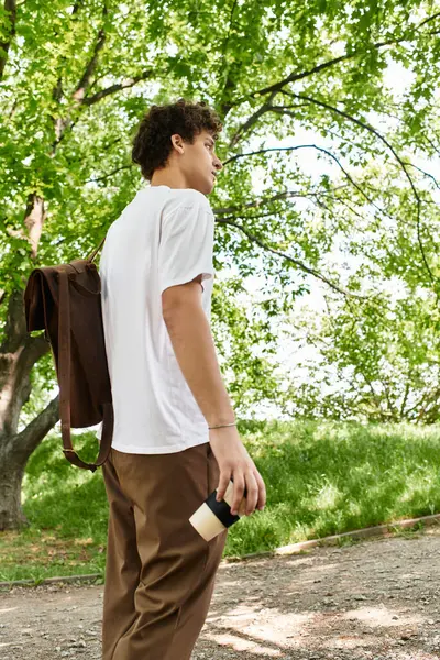 A young man with curly hair casually strolls through a lush park, sipping coffee and relaxing. — Stock Photo