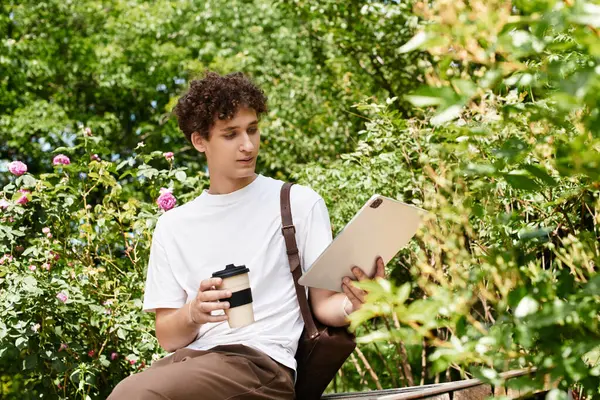A young man relaxes in a green park, reading on a tablet and enjoying a cup of coffee. — Stock Photo