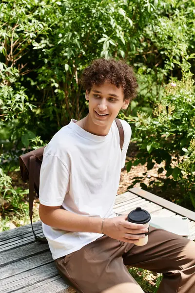 A young man with curly hair sits on a bench, savoring a drink in the lush greenery of summer. — Stock Photo