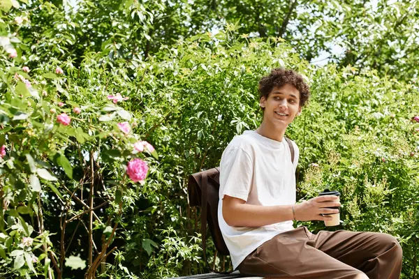 A young man with curly hair relaxes in a green park, savoring his drink among blooming flowers. — Stock Photo