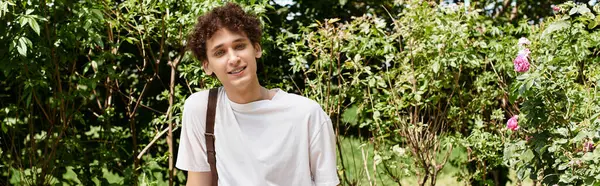 A young man with curly hair smiles while relaxing in a lush summer park filled with greenery, banner — Stock Photo