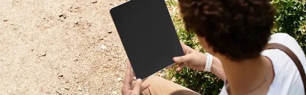 A young man with curly hair relaxes outdoors in a lush summer park while engaging with his tablet, banner — Stock Photo