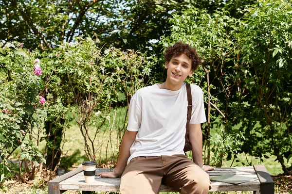 In a green summer park, a young man relaxes on a bench with a coffee, enjoying nature beauty. — Stock Photo