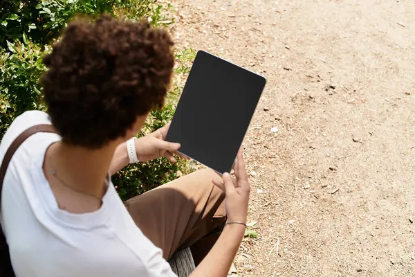 A young man with curly hair relaxes in a sunny park while holding a tablet, enjoying nature. — Stock Photo