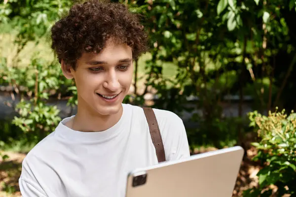 A young man with curly hair is focused on a tablet in a vibrant summer park, smiling. — Stock Photo