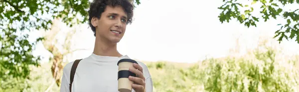 A young man with curly hair relaxes in a lush park, holding a cup of coffee and smiling warmly, banner — Stock Photo