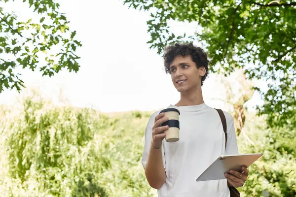 A young man with curly hair relaxes with coffee and his tablet in a sunny park. — Stock Photo