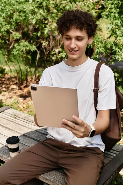 Curly-haired young man sits in a green park, happily using his tablet and sipping coffee. — Stock Photo