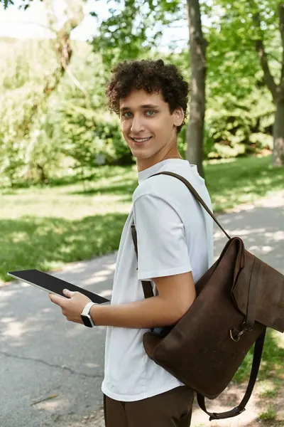 A young man with curly hair relaxes in a vibrant summer park, engaging with a tablet. — Stock Photo