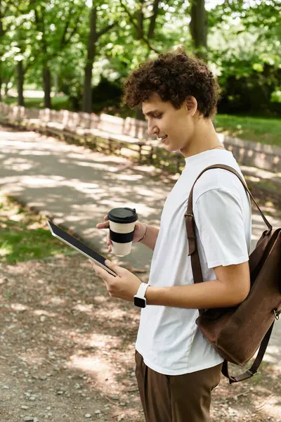 A young man with curly hair relaxes in a vibrant summer park, reading on a tablet. — Stock Photo