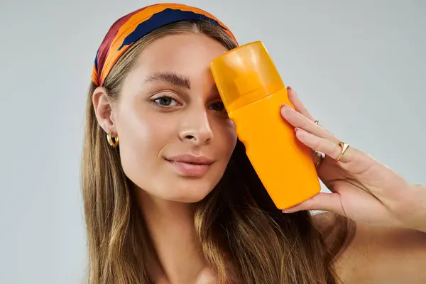 Young woman with a headscarf holds a bright orange bottle, showcasing summer fashion in a studio. — Stock Photo