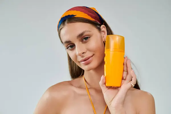 A confident young woman with tan skin poses in a summer outfit, holding a bright orange sunscreen. — Stock Photo