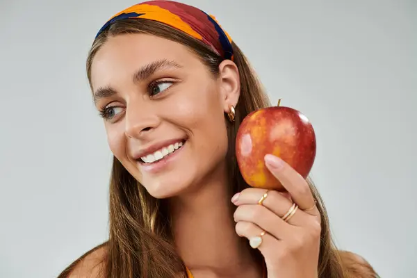 Young woman with tan skin holds a red apple while smiling in a stylish summer outfit. — Stock Photo