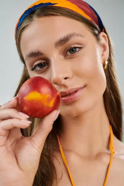 A young woman with tan skin and a vibrant headscarf holds a fresh peach, exuding summer beauty. — Stock Photo