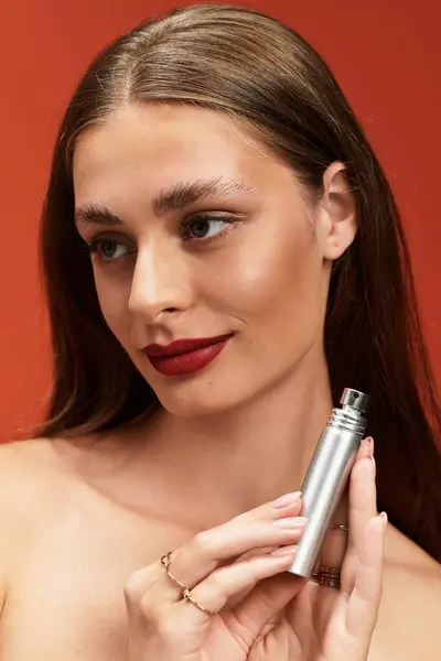 Young woman with glowing skin holds a sleek fragrance bottle against a vibrant background. — Stock Photo