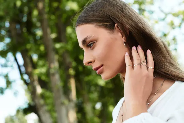 Captivating young woman with long hair gently touching her jewelry amidst lush greenery. — Stock Photo