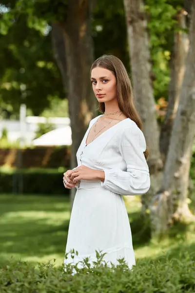 A beautiful young woman poses elegantly in a white dress amidst lush greenery on a sunny day. — Stock Photo