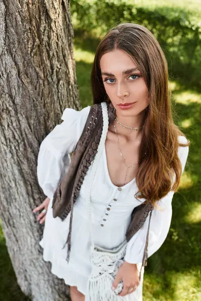 A young woman stands by a tree, highlighting her long hair and stylish outfit in sunlight. — Stock Photo