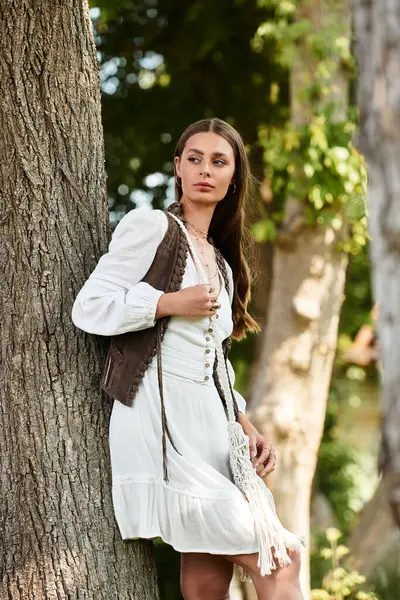 A graceful young woman in a white outfit stands by a tree in soft sunlight. — Stock Photo