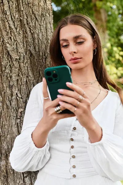 Молодая красивая женщина стоит рядом с деревом, помолвленная со своим смартфоном в солнечный день. — стоковое фото