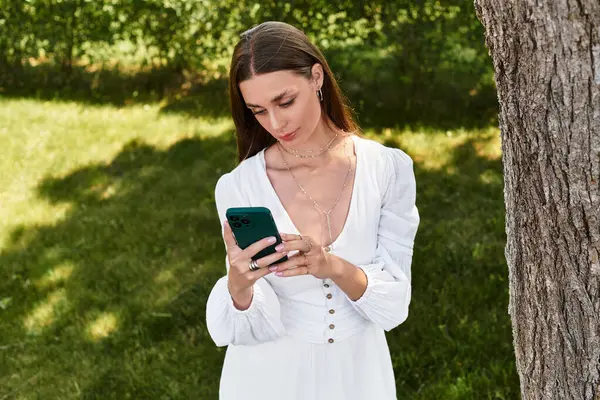 A young beautiful woman stands near a tree, absorbed in her smartphone amidst vibrant greenery. — Stock Photo